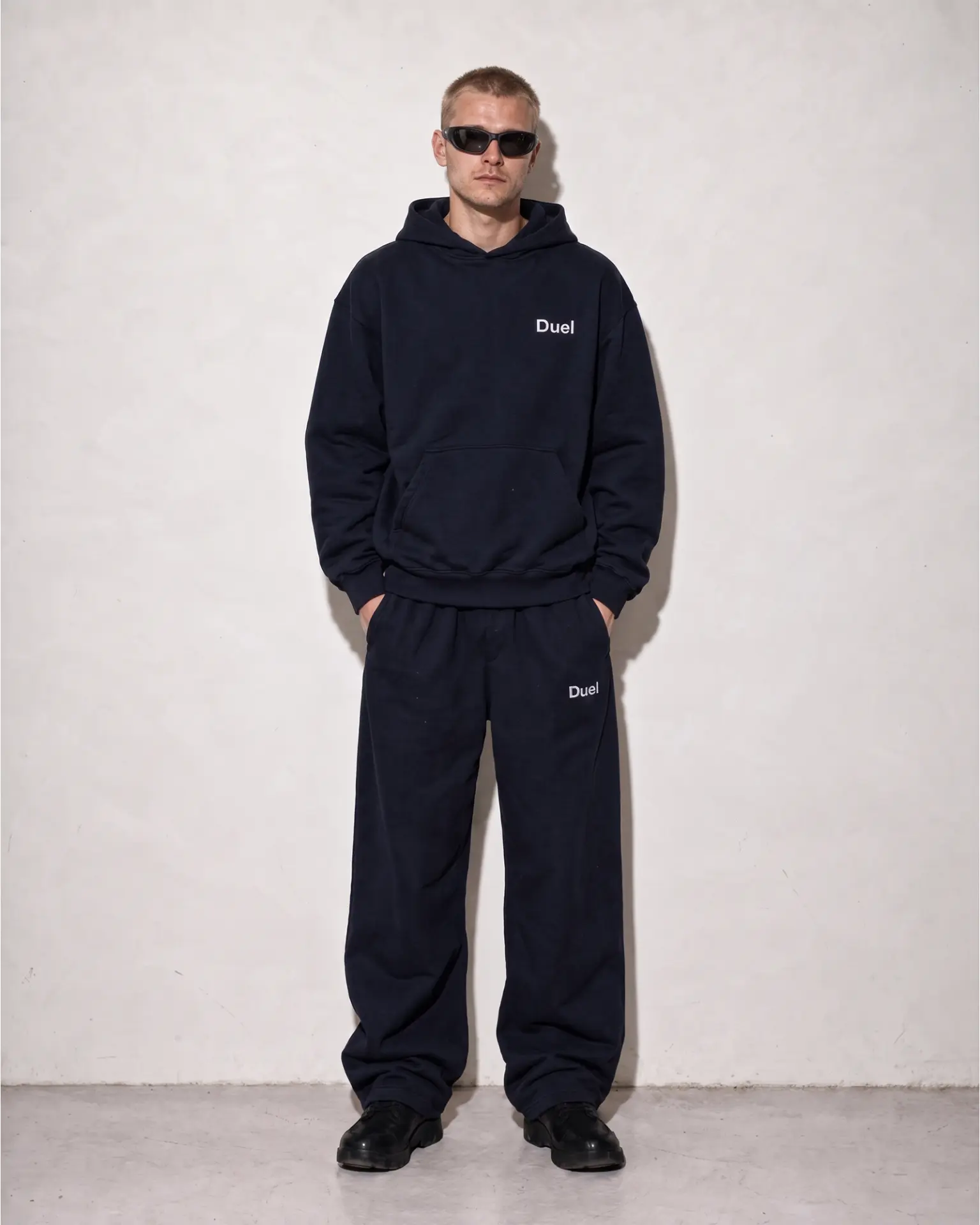 Campaign photograph: model in sunglasses wearing a navy Duel hoodie and matching trousers against a white studio wall
